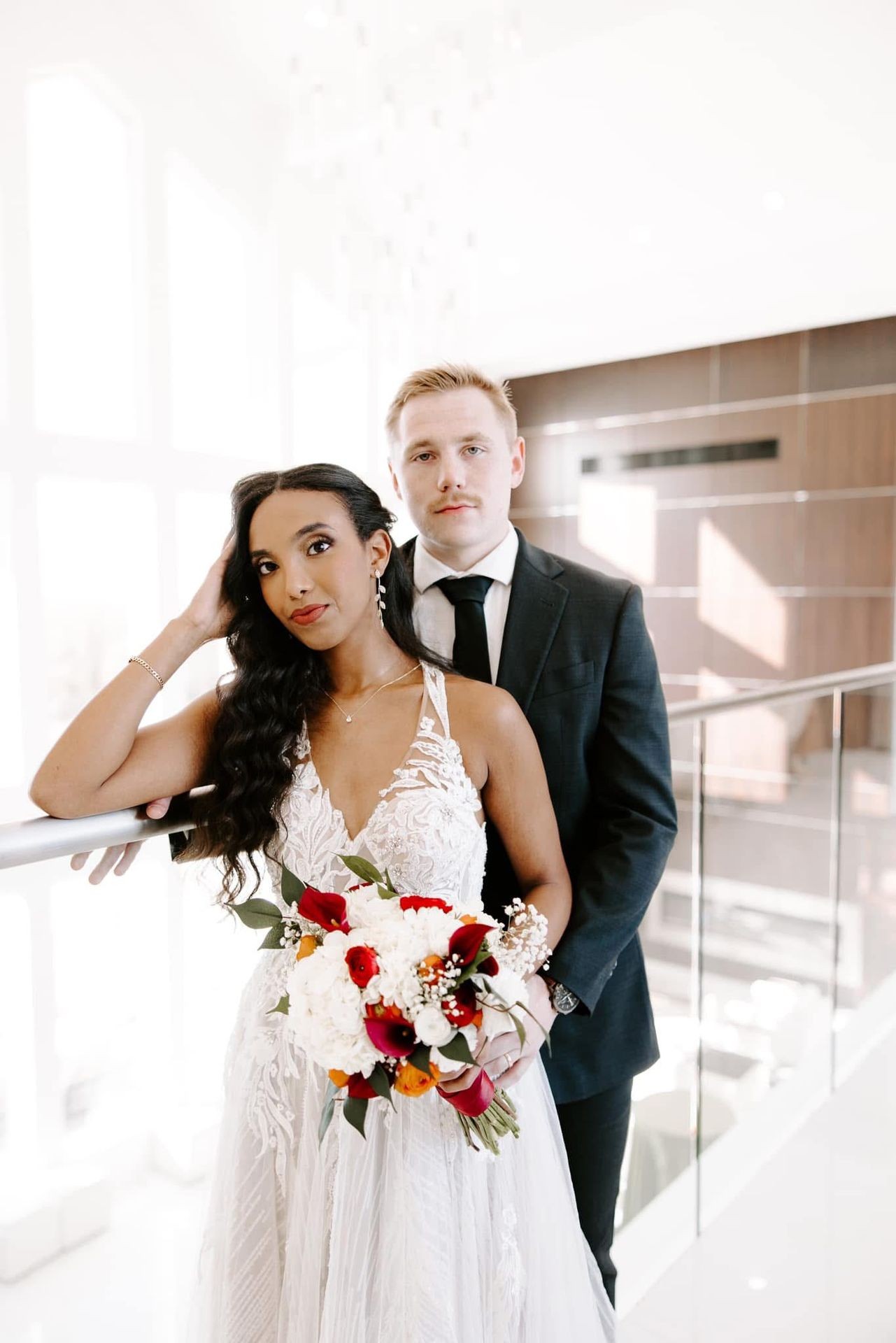 Bride with bouquet and groom in formal attire posing indoors with natural light.