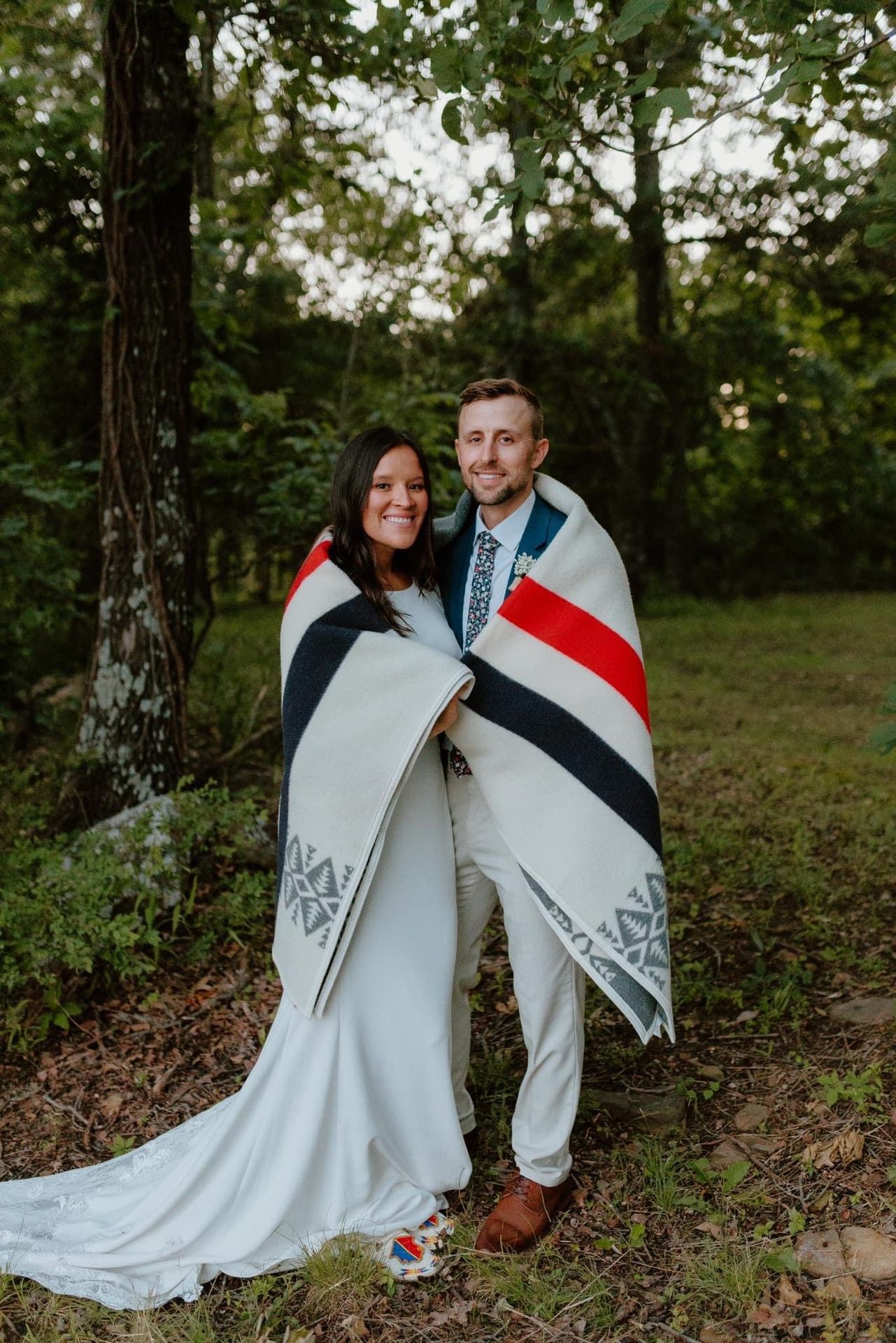 Bride and groom wrapped in a patterned blanket standing outdoors in a forest setting.