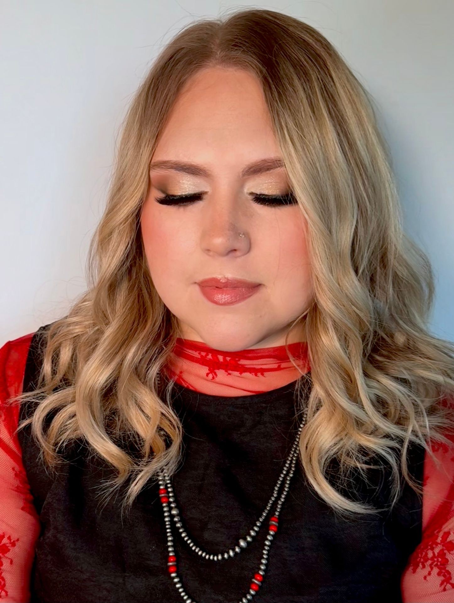 Woman with closed eyes, blonde wavy hair, wearing a red top and necklace.