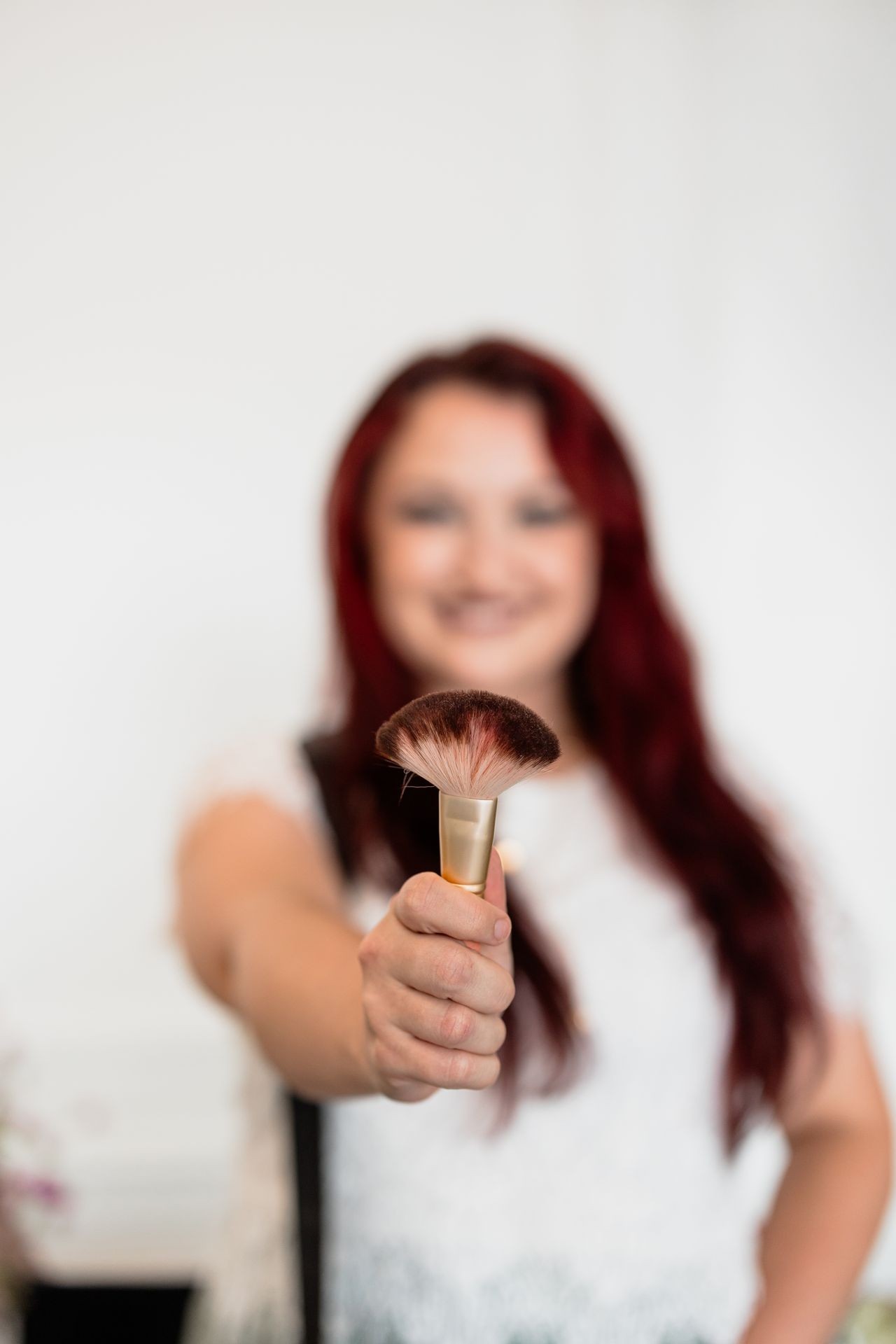 Person with long, red hair extending a makeup brush towards the camera, wearing a white shirt.