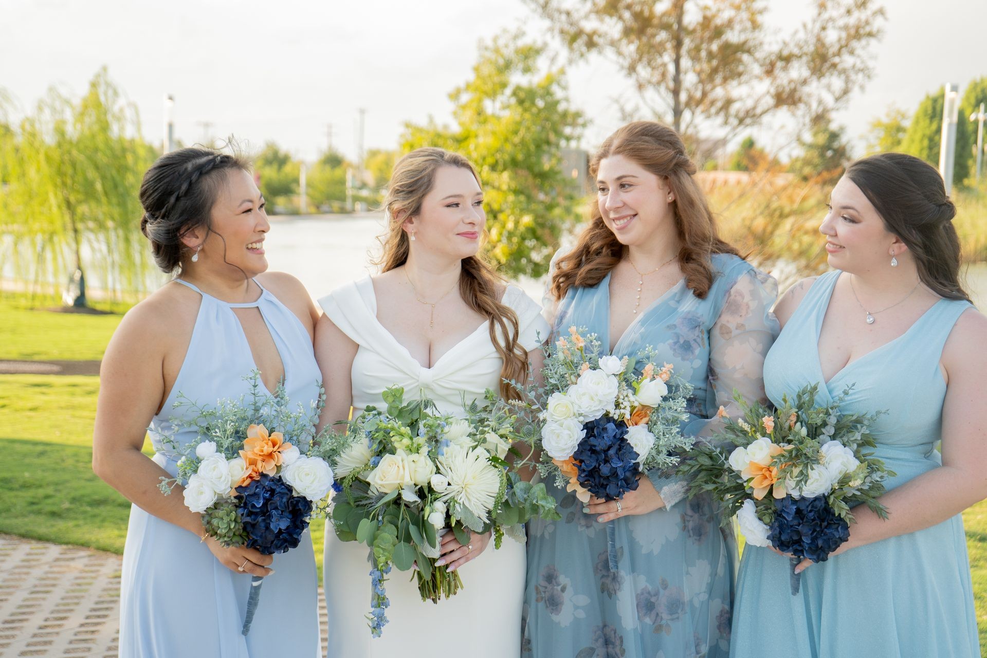 Four women in elegant dresses holding floral bouquets, smiling outdoors on a sunny day.